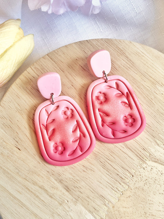 Statement Floral Polymer Clay Earrings from silverstar.nz feature a unique design with pink floral and leaf patterns on a rectangular shape with rounded edges, set against a wooden surface. These handmade earrings showcase darker pink flower and leaf designs paired with a lighter pink top piece.