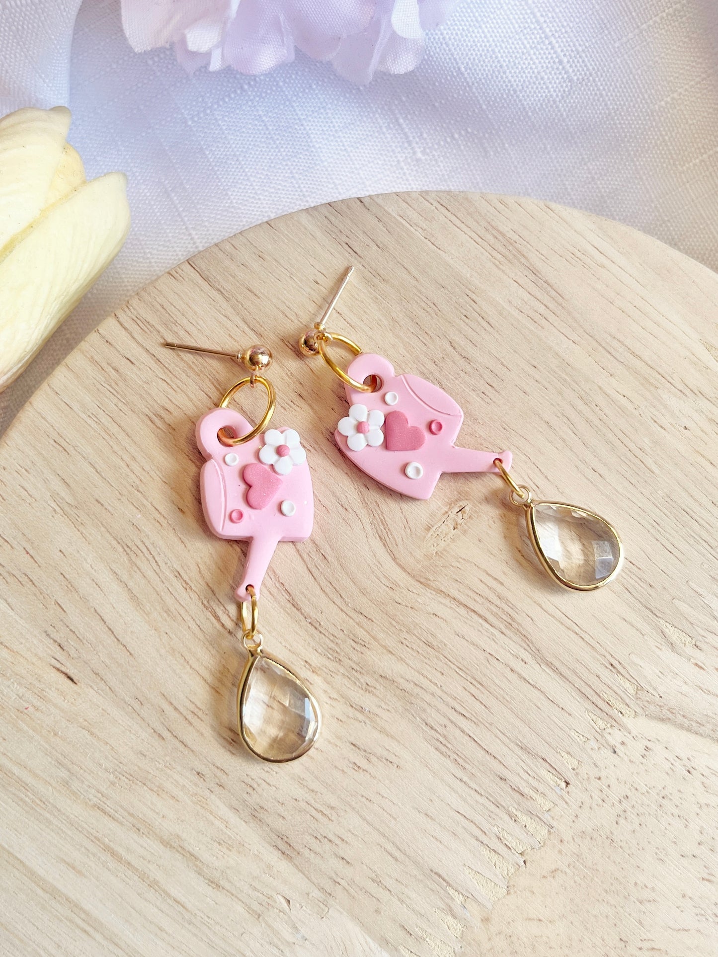 The Love Watering Can Polymer Clay Earrings by silverstar.nz are whimsical, handmade pieces featuring pink watering cans with flowers and gold studs. Clear teardrop beads dangle from stainless steel hooks, and the earrings are shown resting on wood next to light-colored flowers.