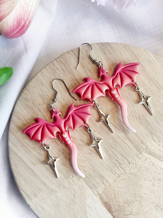 The Valentines Dragon Polymer Clay Earrings by silverstar.nz, featuring pink dragon designs with dangling star charms, rest on a round wooden surface. In the background, a soft pink flower with subtle green leaves accentuates the artistry of these handmade earrings.