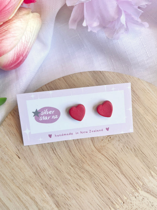 Valentines Heart Polymer Clay Stud Earrings feature a funky design on a card with silverstar.nz branding and handmade in New Zealand text, resting amidst pink and white flowers on wood, highlighting their vibrant allure.