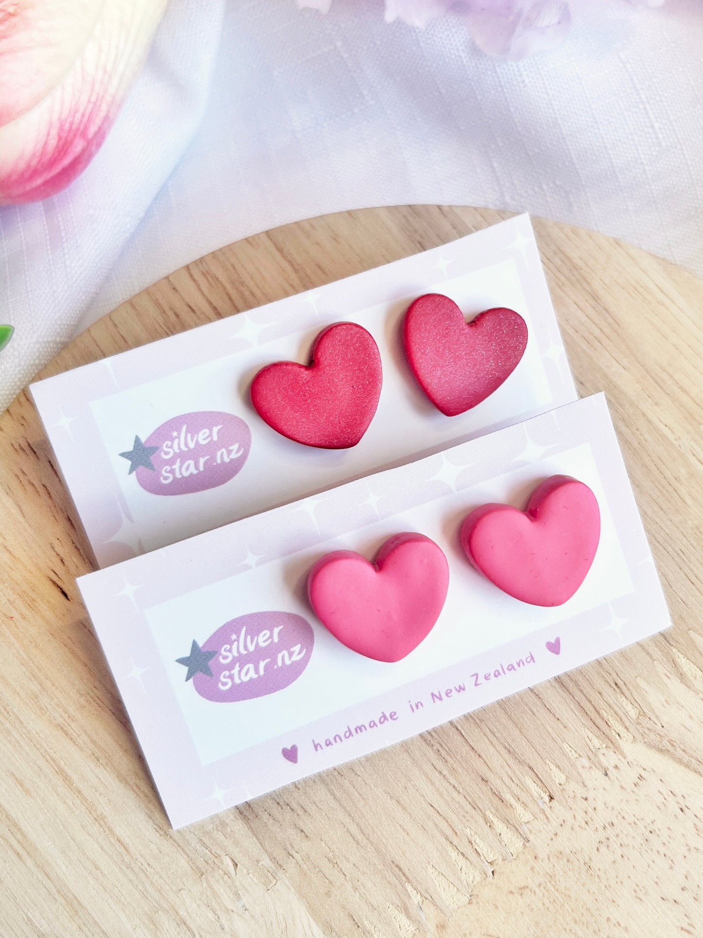 Two pairs of heart-shaped earrings, one red and one pink, on white silverstar.nz cards. These Valentines Heart Polymer Clay Stud Earrings feature handmade clay hearts on stainless steel hooks, set against soft pink and white fabric over light wood.