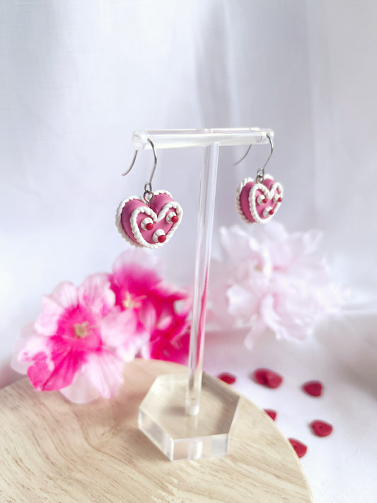 The Vintage Heart Cake Polymer Clay Earrings by silverstar.nz hang on a transparent stand, featuring pink heart-shaped designs with white and red details. They include stainless steel hooks and are displayed on a wooden base adorned with pink flowers and heart decorations in the background.