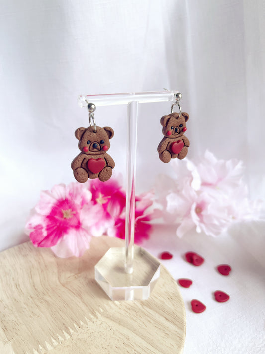 The Love Teddy Bear Polymer Clay Earrings by silverstar.nz feature bear shapes with red hearts, crafted from polymer clay and hanging from stainless steel hooks. Theyre displayed on a clear stand with pink and white flowers, plus small red heart decorations scattered on a wooden surface in the background.