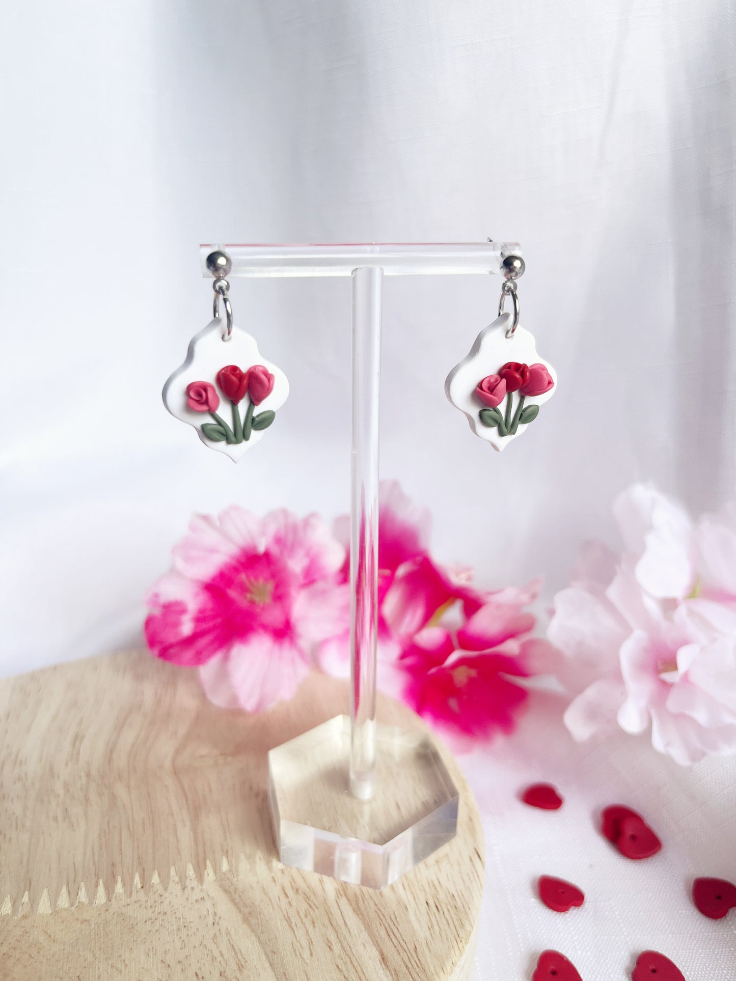 The Tulip Floral Polymer Clay Earrings by silverstar.nz dangle elegantly from a clear stand, showcasing their handmade design with stainless steel hooks amidst white and pink flowers on a light wooden surface.