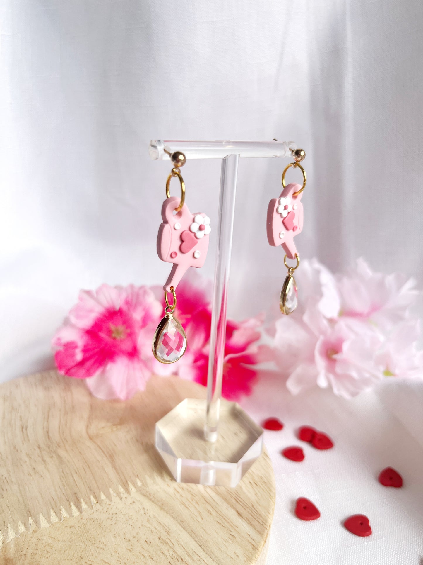 The Love Watering Can Polymer Clay Earrings by silverstar.nz elegantly dangle on a clear display stand, with a backdrop of pink flowers and red heart decorations on a wooden surface with white fabric.