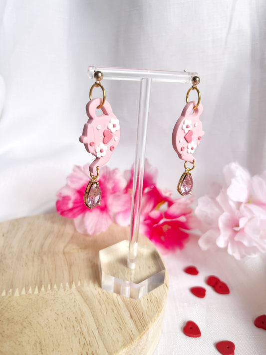 Displayed on a clear stand, the Love Tea Pot Polymer Clay Earrings from silverstar.nz feature teardrop-shaped pink crystals, complemented by pink flowers and red heart decorations on a light wooden surface.