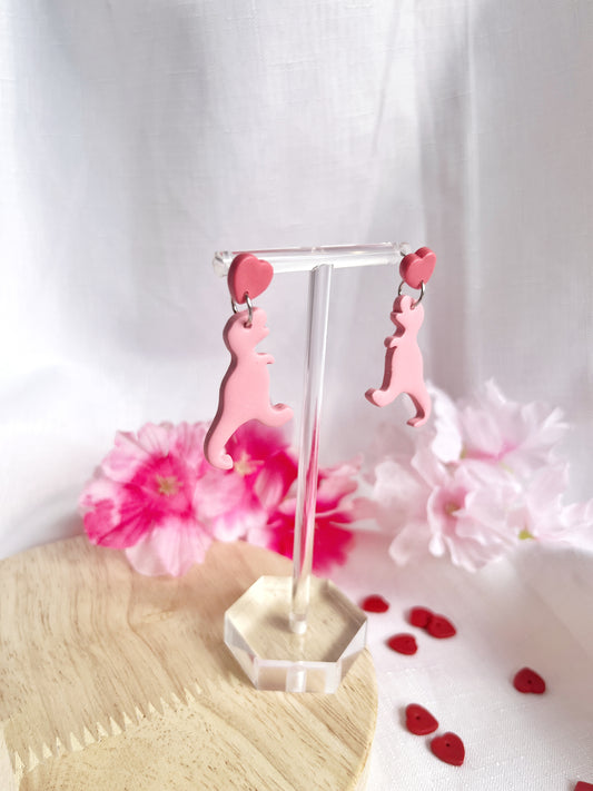 The Heart Dino Polymer Clay Earrings from silverstar.nz blend a unique design with playful charm. These pink dinosaur-shaped earrings feature heart-shaped studs and are elegantly displayed against flowers and small red hearts on fabric and a wooden surface.