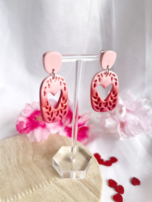 The Leafy Heart Dangle Polymer Clay Earrings by silverstar.nz, featuring intricate floral cutouts and stainless steel hooks, are displayed on a stand against pink flowers and scattered red hearts on a wooden surface.
