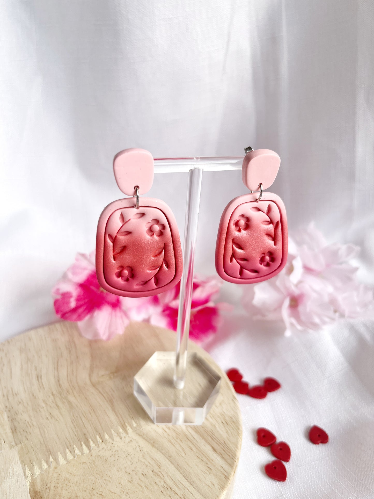 The Statement Floral Polymer Clay Earrings by silverstar.nz showcase a pink floral design with a rounded triangular top and larger rectangular piece, displayed on a clear stand over wood with pink flowers and heart accents nearby.