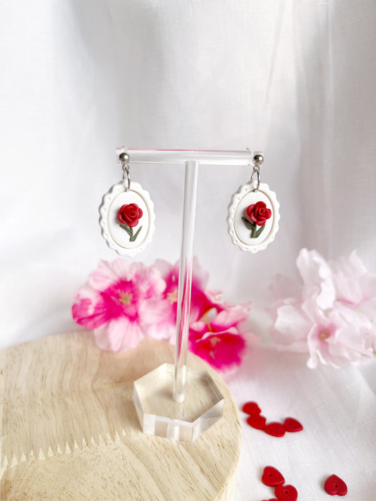 Framed Rose Polymer Clay Earrings by silverstar.nz feature elegant handmade red roses on white ovals. Soft pink flowers and heart decorations on a light wood base, with durable stainless steel hooks, complete this graceful set perched on a clear stand.