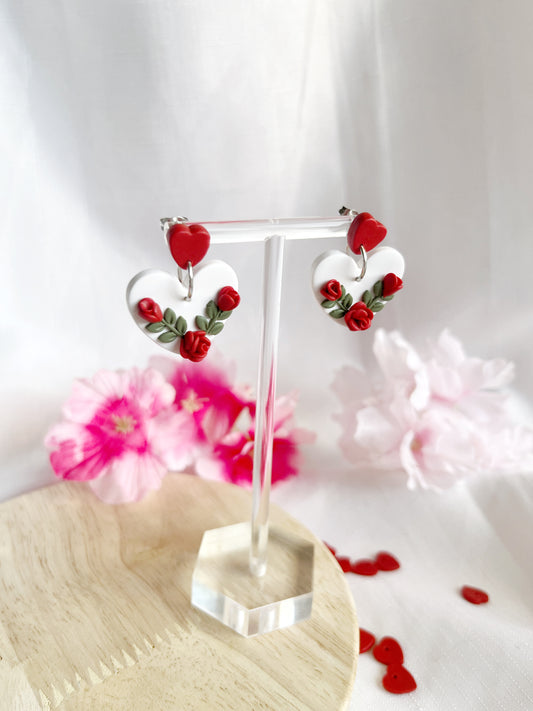 Heart Rose Floral Polymer Clay Earrings by silverstar.nz are showcased on a clear stand with pink flowers and small red heart decorations in the background, set against a light wood surface, highlighting their funky design.