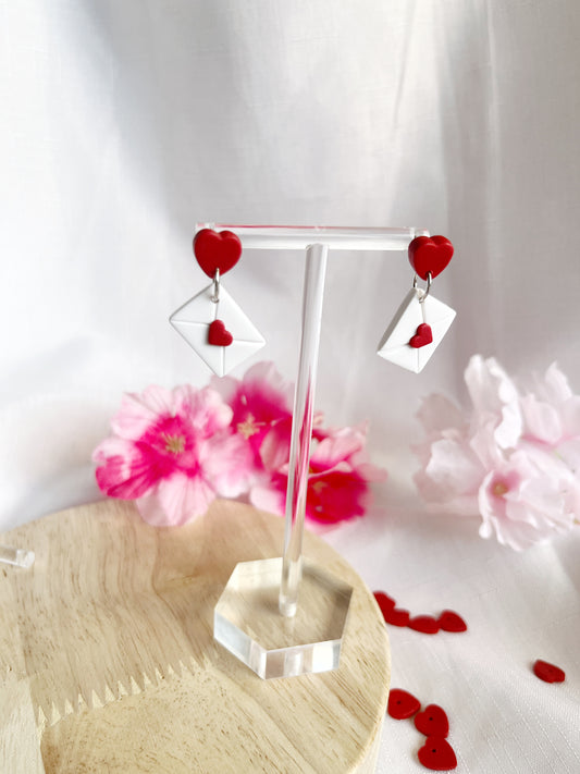 Valentines Envelope Polymer Clay Earrings by silverstar.nz elegantly hang on a clear stand, set against a background of pink flowers and scattered red hearts on a wooden surface, highlighting the charm of these handmade red heart-shaped and white envelope earrings.