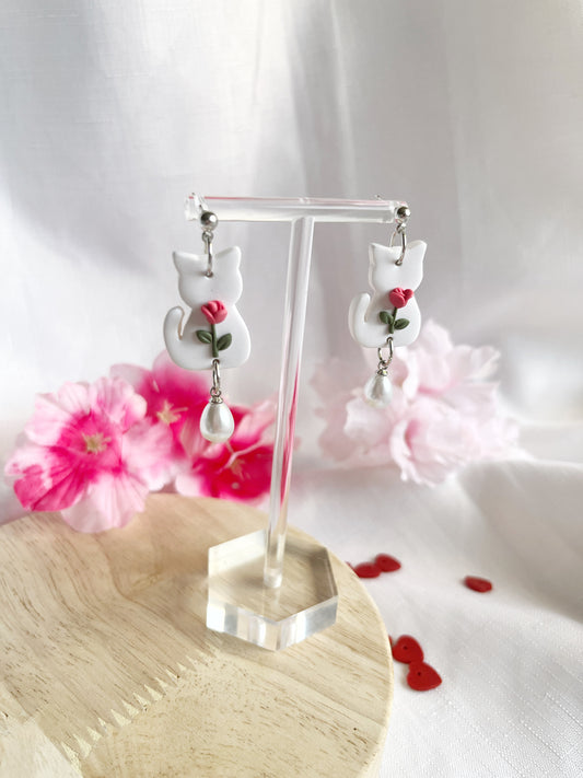 The White Tulip Cat Polymer Clay Earrings by silverstar.nz, handcrafted with stainless steel hooks, feature red roses and pearl drops. They are displayed on a transparent stand with a white fabric backdrop, pink flowers, and scattered red heart decorations on wood.