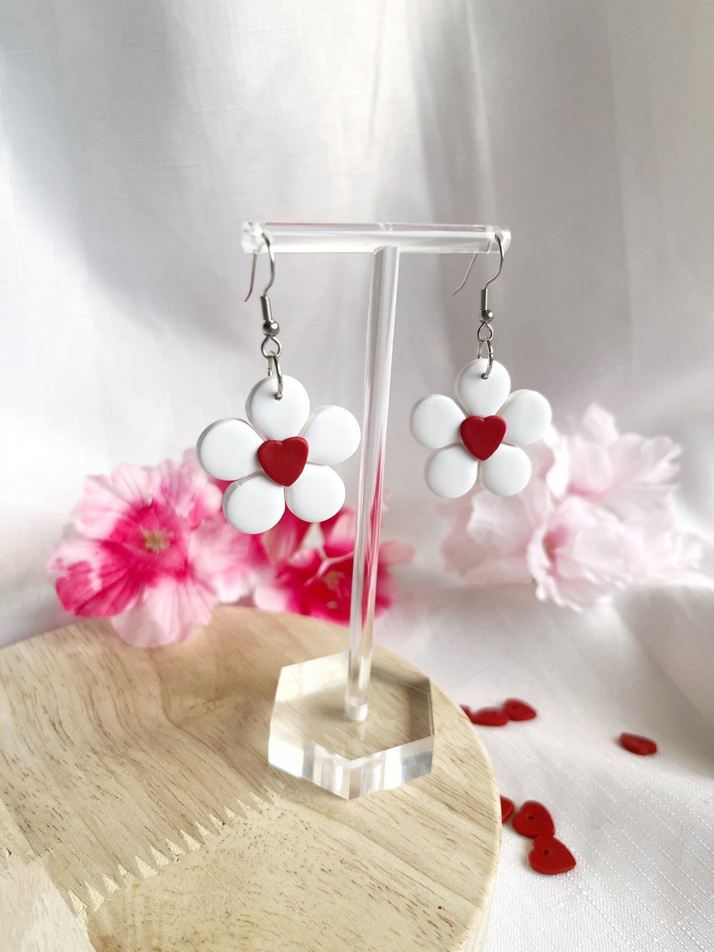 The Daisy Hearts Polymer Clay Earrings by silverstar.nz hang on a clear stand, each featuring white petals and a central red heart. Made with quality stainless steel hooks, theyre set against pink flowers and red heart confetti on a wooden surface.