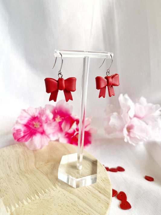 Red Bow Polymer Clay Earrings by silverstar.nz elegantly rest on a wooden surface with stainless steel hooks. Soft-focus pink flowers and scattered red heart-shaped confetti in the background enhance the romantic ambiance, highlighting these handmade earrings.