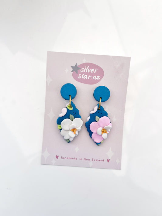 Blue Floral Polymer Clay Earrings by silverstar.nz boast blue and white floral designs with pink accents on a branded card stating handmade in New Zealand. These circular studs feature small flowers with orange centers.