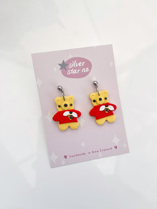 The Honey Bear Polymer Clay Earrings feature teddy bear faces with red capes on stainless steel hooks. Displayed on a card from silverstar.nz, they are crafted from yellow polymer clay and set against a pink starry background labeled handmade in New Zealand.