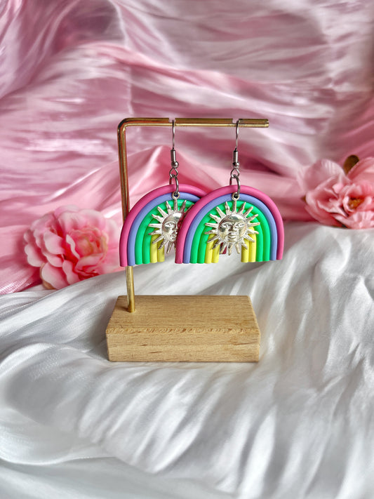 The Rainbow Arch Polymer Clay Earrings by silverstar.nz feature a colorful design with sun charms. Displayed on a gold stand against silky pink and white fabric with pink flowers, these handmade earrings highlight creativity and charm.