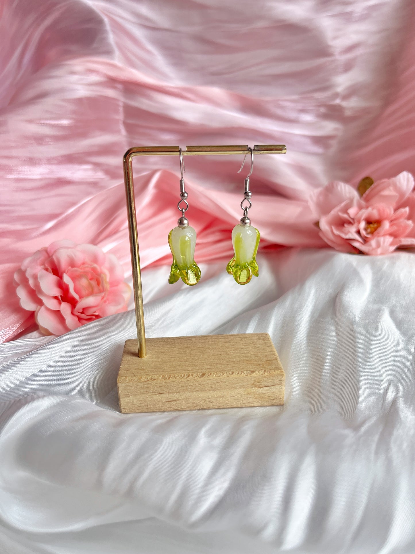 Bok Choy Glass Bead Earrings by silverstar.nz, crafted with handmade flower-shaped glass beads on stainless steel hooks, hang from a gold stand placed on a wooden base with pink silk fabric and soft pink flowers as the backdrop.