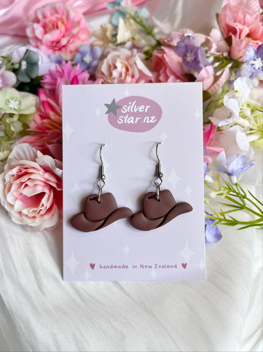 The Cowboy Hat Polymer Clay Earrings from silverstar.nz feature elegant brown cowboy hat designs crafted from polymer clay, dangling from stainless steel hooks on a card labeled handmade in New Zealand, surrounded by pink, purple, and white flowers.