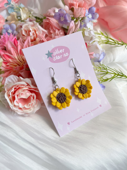 The Sunflower Polymer Clay Earrings by silverstar.nz are presented on a pink card adorned with pink and purple flowers. They rest on a soft white fabric backdrop, enhancing the romantic vibe of these handmade earrings with their playful, funky design.