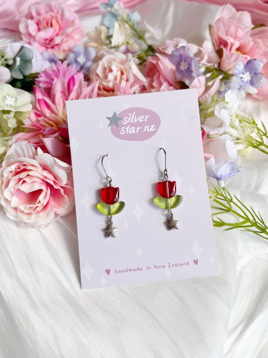 The Tulip Bead Earrings by silverstar.nz are unique handmade pieces shaped like red and green flowers with silver star accents. They feature stainless steel hooks and are displayed on a card with pink and purple floral fabric, reading silver star.nz and handmade in New Zealand.