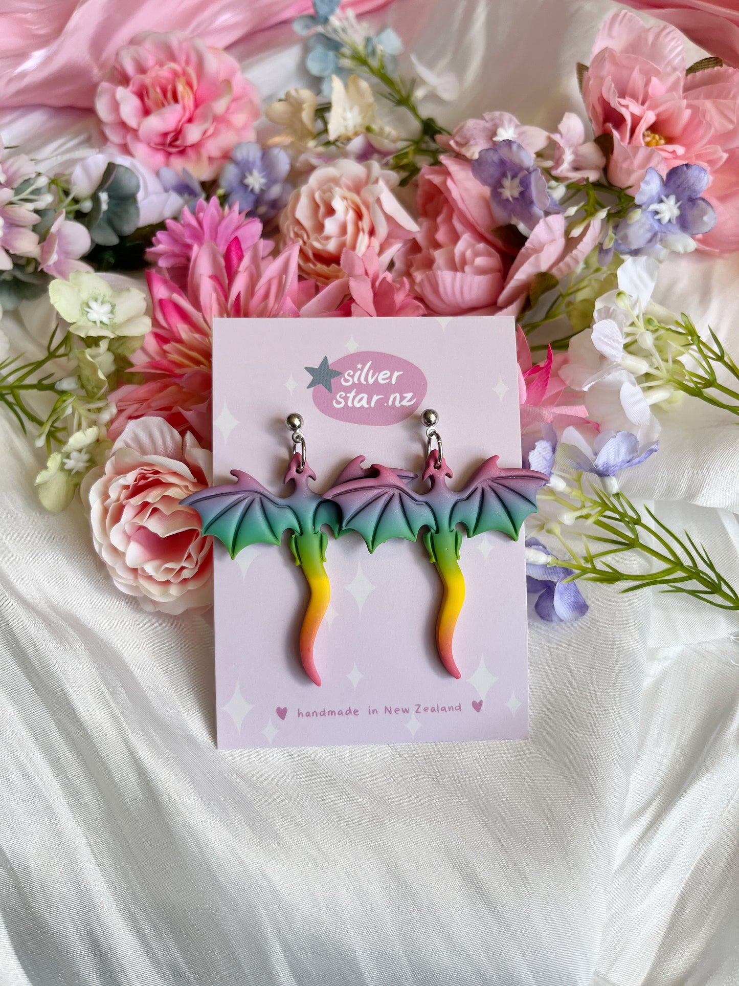 The Rainbow Dragon Polymer Clay Earrings feature pink, green, and yellow hues and dangle from stainless steel hooks. Displayed on a pink card labeled silverstar.nz, they rest among pink and purple flowers on silky white fabric.