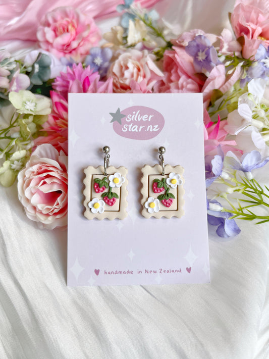 Strawberry Stamp earrings feature a unique white, pink, and green floral design crafted from polymer clay on a silverstar.nz card, surrounded by colorful flowers on a white fabric background.