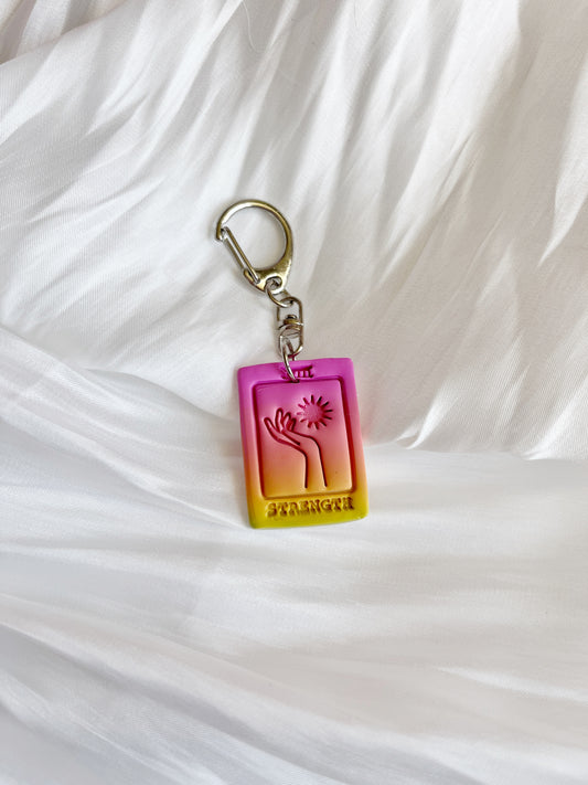A Tarot Card Polymer Clay Keyring by silverstar.nz shows a pink to yellow gradient on a white, textured fabric. Featuring an embossed hand reaching for a sun with STRENGTH, it’s the perfect artisanal piece for those who appreciate unique designs and handmade charm.