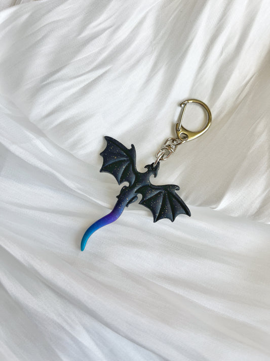The Midnight Blue Dragon Keyring by silverstar.nz features a small black dragon figure with shiny details and a gradient tail from purple to blue, set against a white fabric background. This unique design is handmade from polymer clay, making it an ideal gift.