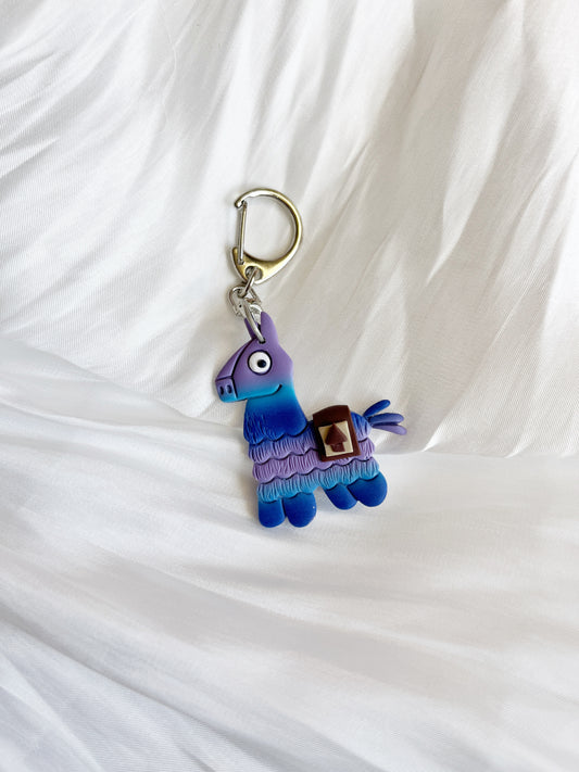 The Fortnite Llama Polymer Clay Keyring by silverstar.nz features a unique blue and purple llama design with a saddle against a crumpled white fabric background. It includes a silver-colored metal clasp, making it an ideal handmade gift.