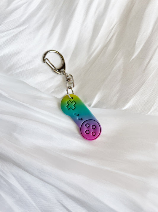 The silverstar.nz Retro Game Controller Polymer Clay Keyring is uniquely designed in the shape of a retro controller, features a durable polymer clay build, and displays a gradient of purple, blue, and green hues against a soft white fabric background.
