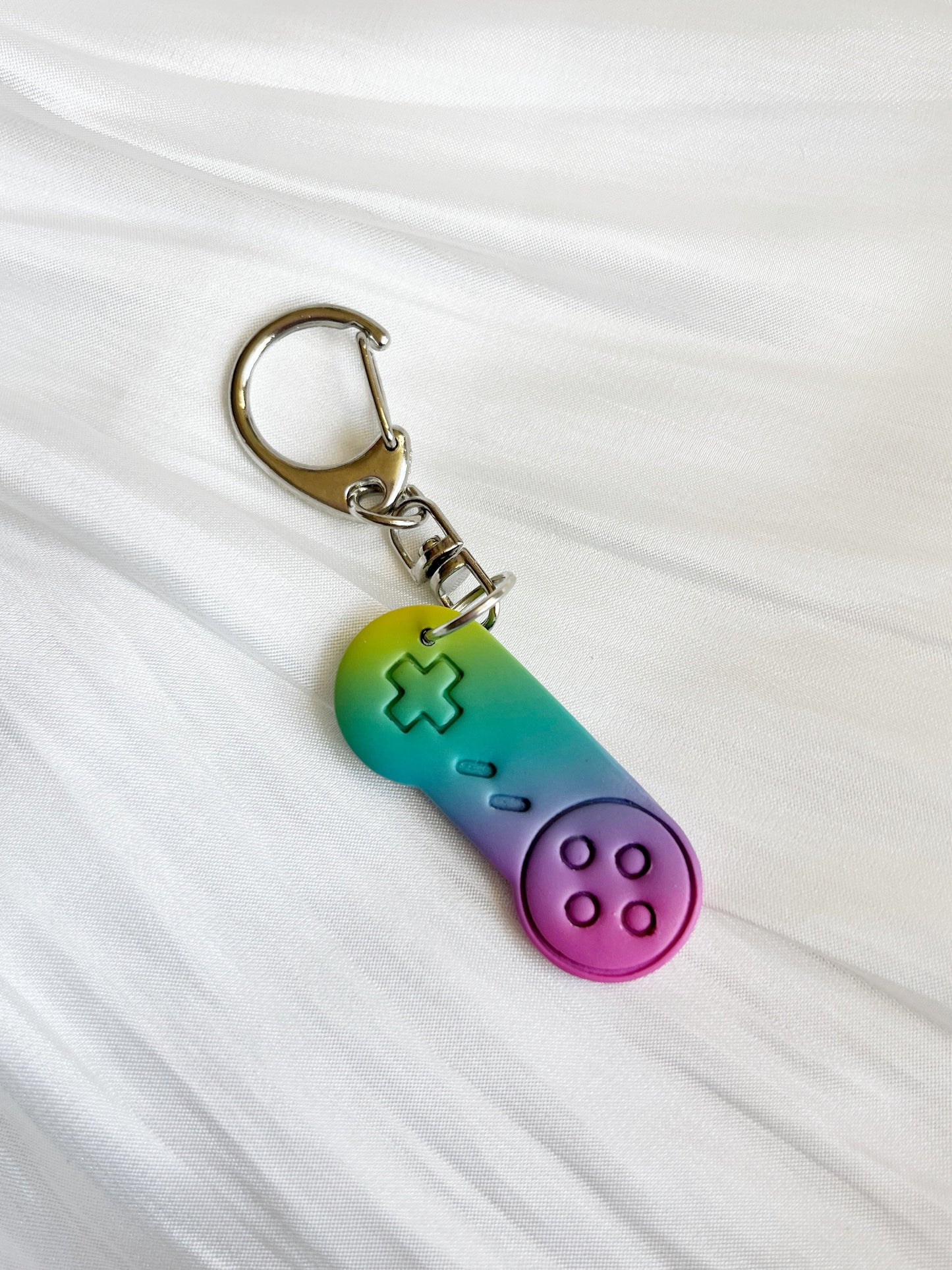 The Retro Game Controller Polymer Clay Keyring by silverstar.nz showcases a unique handmade game controller charm with a green, blue, and pink gradient. It features four round buttons and a cross-shaped directional pad, beautifully displayed on a soft white fabric background.