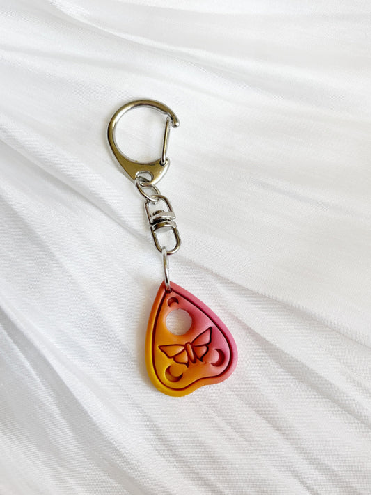The Ouija Planchette Polymer Clay Keyring from silverstar.nz features a heart-shaped red and yellow pendant with an engraved butterfly, attached to a silver keyring. Handmade in New Zealand, it rests on textured white fabric, highlighting its artisanal charm.