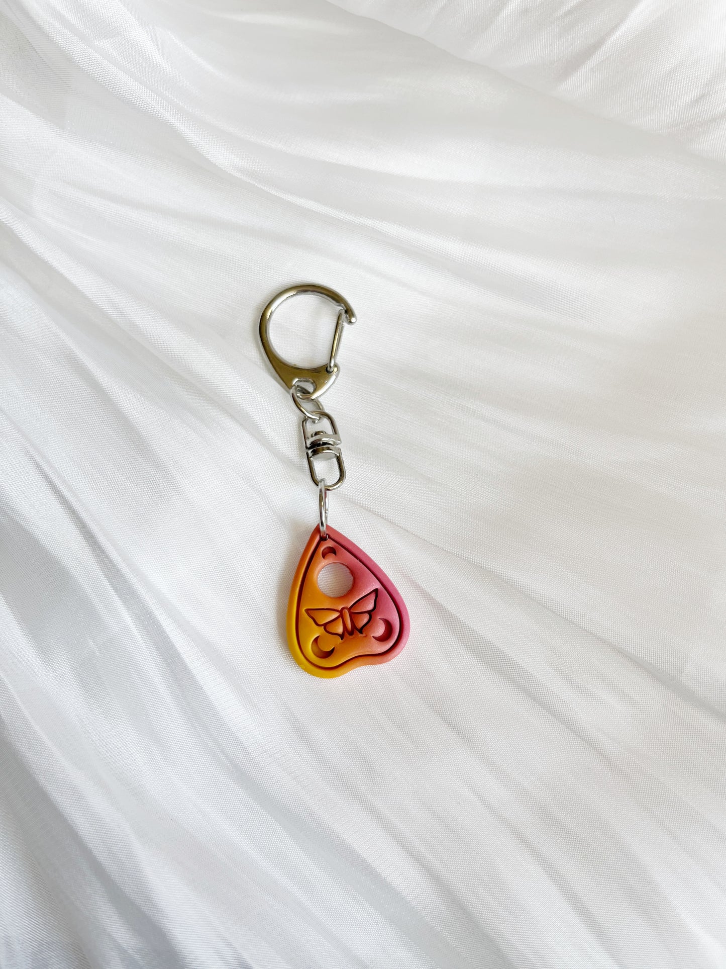 The Ouija Planchette Polymer Clay Keyring by silverstar.nz, handmade in New Zealand, features a unique butterfly design with a yellow-to-pink gradient and is attached to a silver clip, elegantly displayed on soft white fabric.