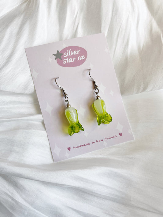 A card labeled silverstar.nz showcases the Bok Choy Glass Bead Earrings, handmade in New Zealand. These earrings, resembling green bell peppers, are adorned with stainless steel hooks and rest against a soft white fabric backdrop.