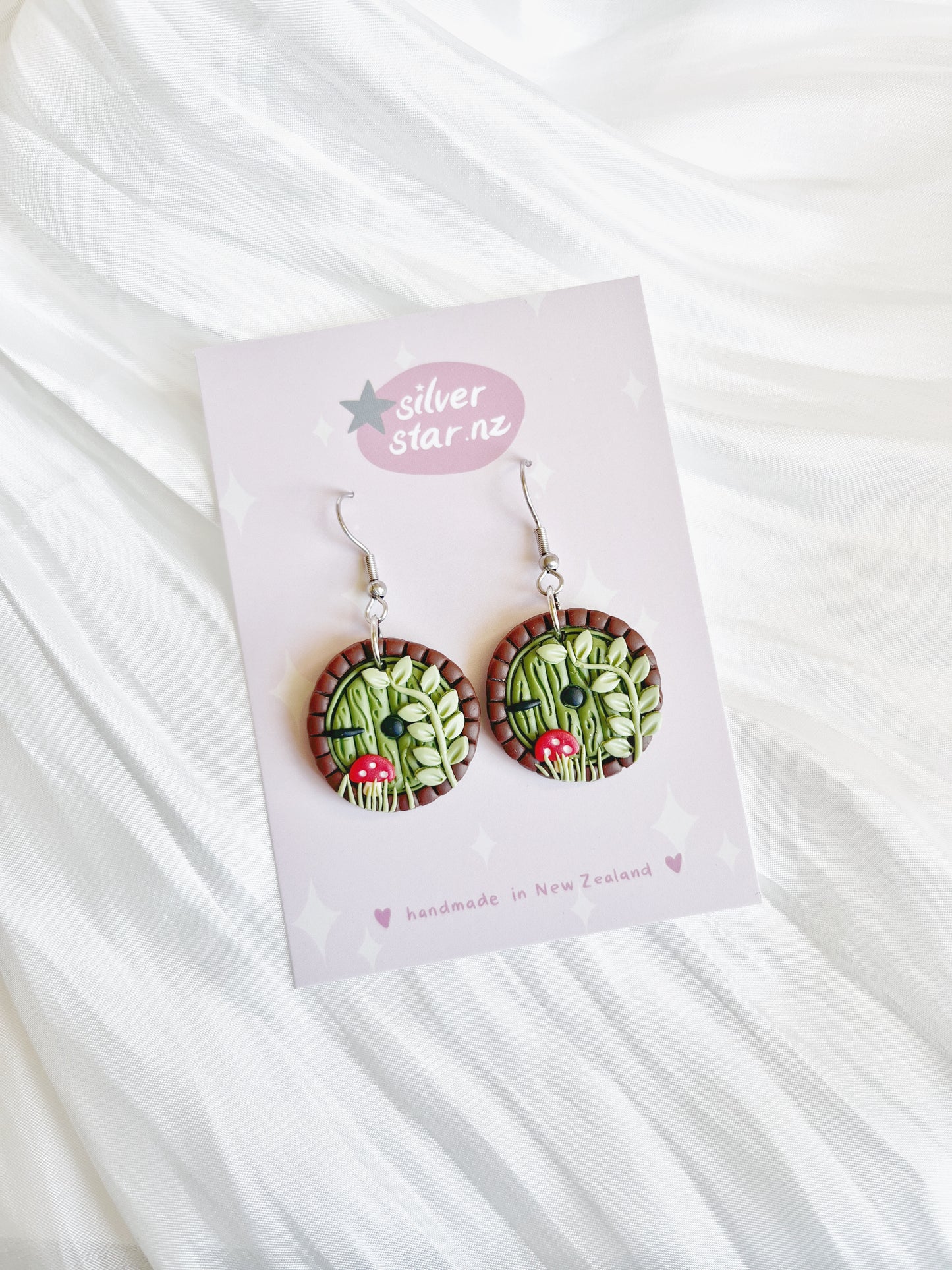 Hobbit Door Polymer Clay Earrings by silverstar.nz feature a round design with green leaves and red fungi accents. Handmade in New Zealand, they are displayed on a white card from silverstar.nz, against a crumpled white fabric background.