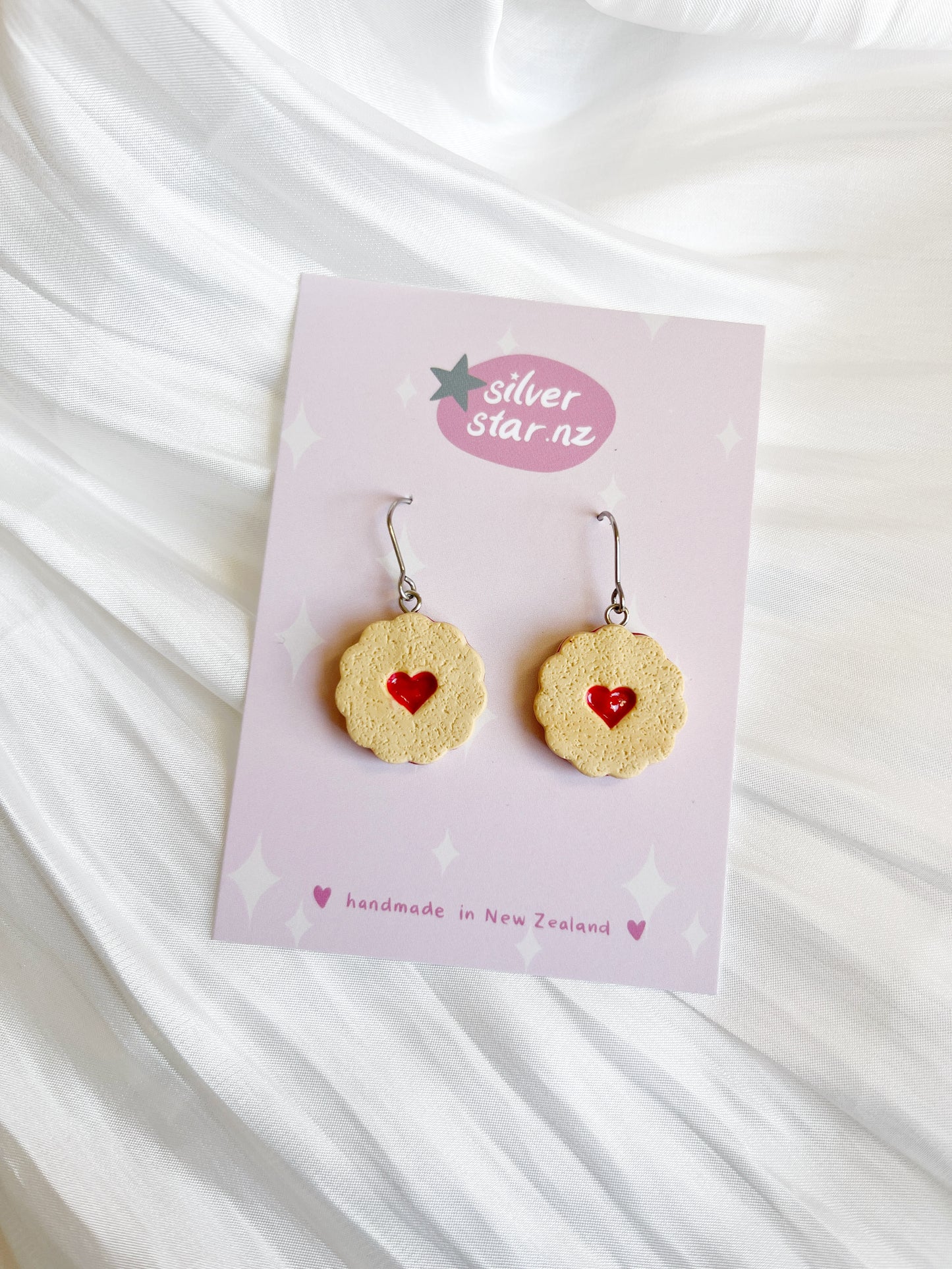 The Shrewsbury Polymer Clay Earrings feature cookie-shaped designs with red heart centers, displayed on a silverstar.nz card. They have stainless steel hooks and are labeled handmade in New Zealand, elegantly placed on a white textured fabric background.