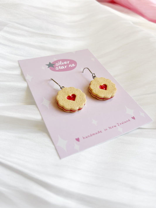 The Shrewsbury Polymer Clay Earrings, crafted to resemble sandwich biscuits with red heart centers, rest on a white fabric background with stainless steel hooks. Displayed on a light pink card labeled silverstar.nz and handmade in New Zealand.