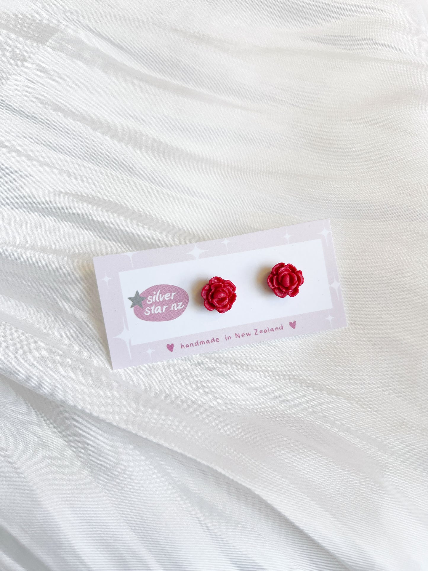 Red rose-shaped polymer clay earrings, labeled as Rose Polymer Clay Stud Earrings by silverstar.nz, sit elegantly on a white card with the handmade in New Zealand text and logo, set against a textured white fabric background.