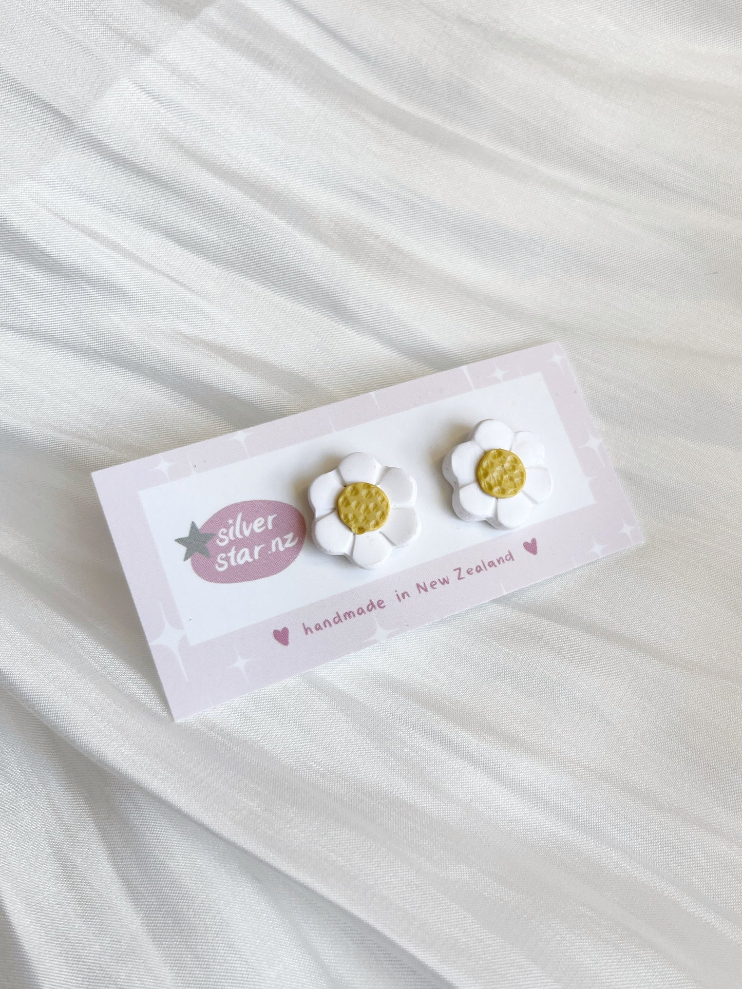 Displayed on a white textured fabric, silverstar.nz presents their White Daisy Polymer Clay Stud Earrings, featuring daisy-shaped earrings with white petals and yellow centers, proudly handmade in New Zealand.
