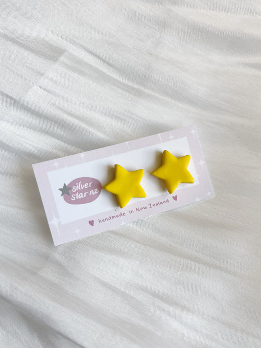 A pair of small, yellow star-shaped handmade polymer clay earrings are shown on a card labeled silverstar.nz with handmade in New Zealand underneath, resting on a textured white fabric background to highlight their unique design.