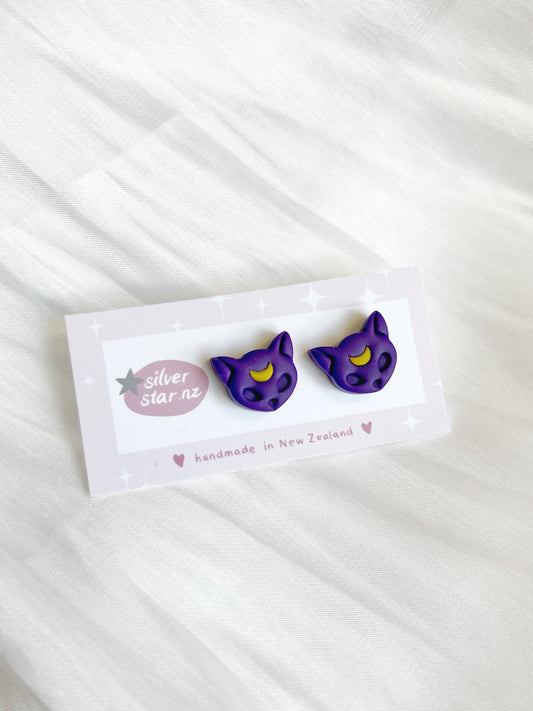 A pair of Luna Cat Polymer Clay Stud Earrings, featuring a unique design with purple cat faces, yellow eyes, and a mischievous smile, are displayed on a white card labeled silverstar.nz and handmade in New Zealand with a heart. The card is set against textured white fabric.