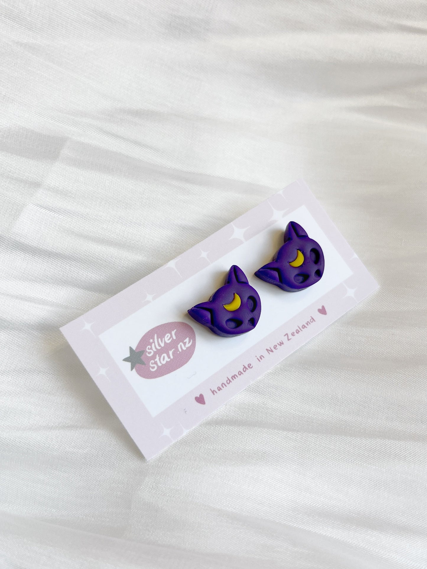 Close-up of Luna Cat polymer clay stud earrings, featuring a purple cat with yellow eyes, on a card displaying the brand silverstar.nz. Resting on soft white fabric, these handmade New Zealand earrings stand out for their unique design.