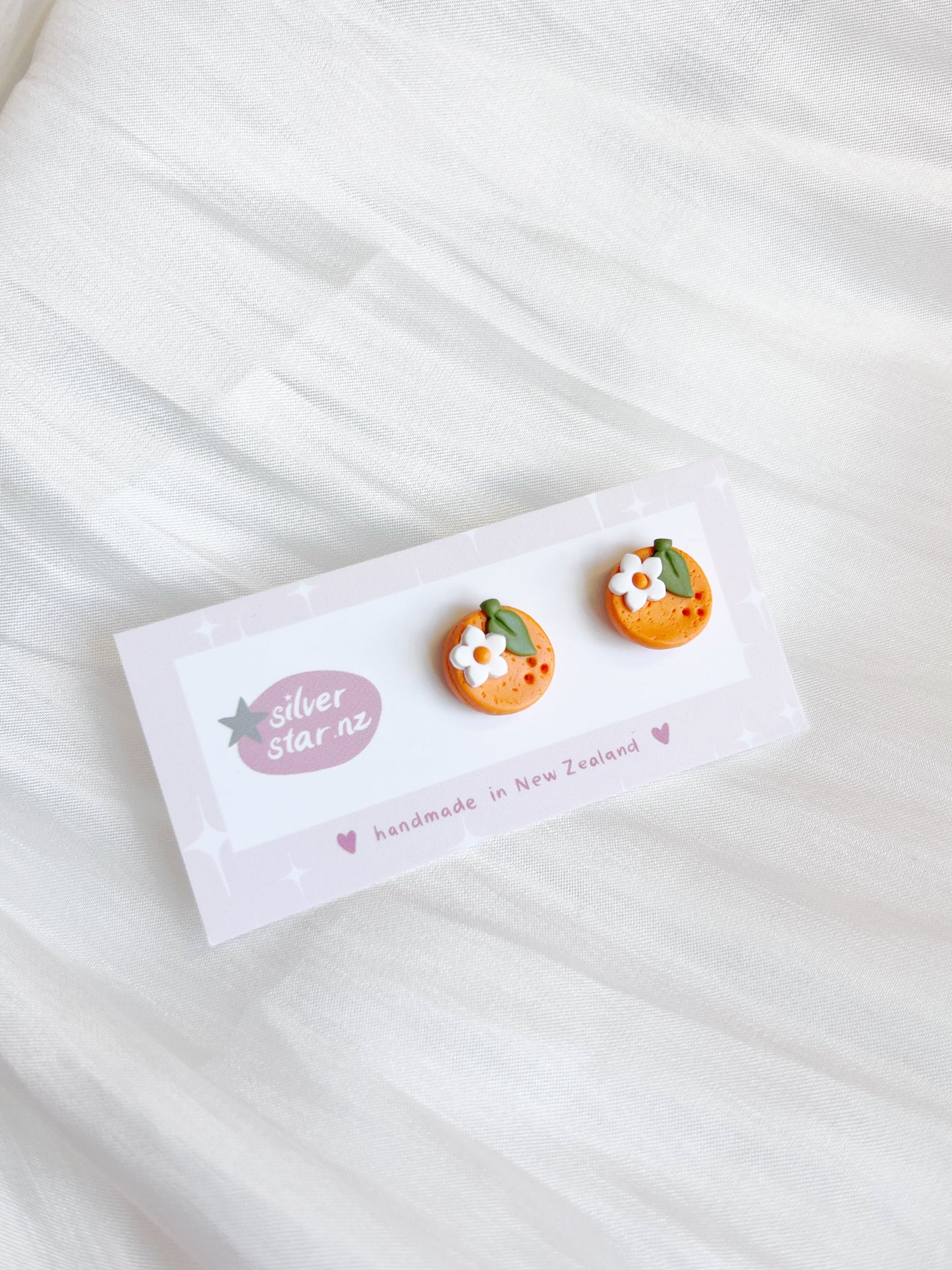 The Orange Blossom Polymer Clay Stud Earrings from silverstar.nz, shaped like oranges with small white flowers, are on a Silver Star NZ card that reads handmade in New Zealand, set against a soft white fabric background.