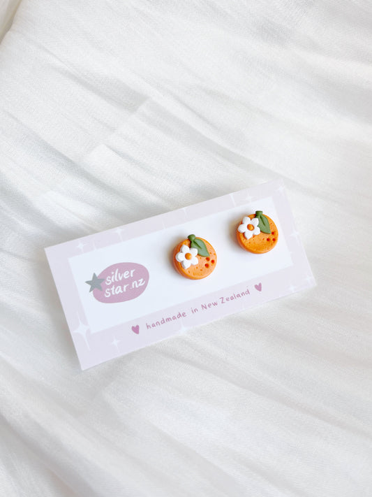 The Orange Blossom Polymer Clay Stud Earrings from silverstar.nz, shaped like oranges with small white flowers, are on a Silver Star NZ card that reads handmade in New Zealand, set against a soft white fabric background.