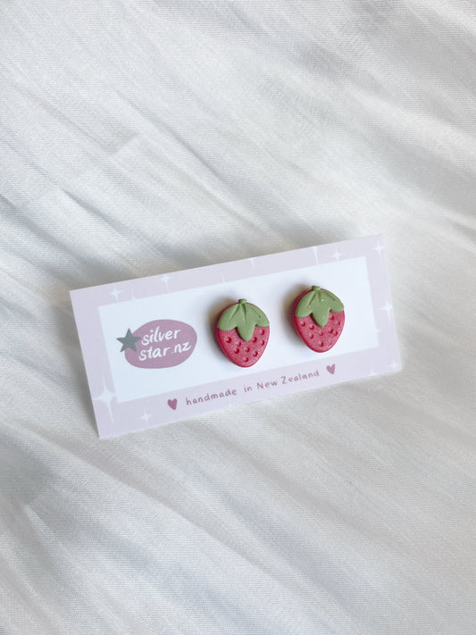 Pink strawberry-shaped polymer clay stud earrings with green leaves rest on a white card labeled silverstar.nz against a textured white fabric background. The card notes they are handmade in New Zealand, featuring durable stainless steel hooks for lasting wear.