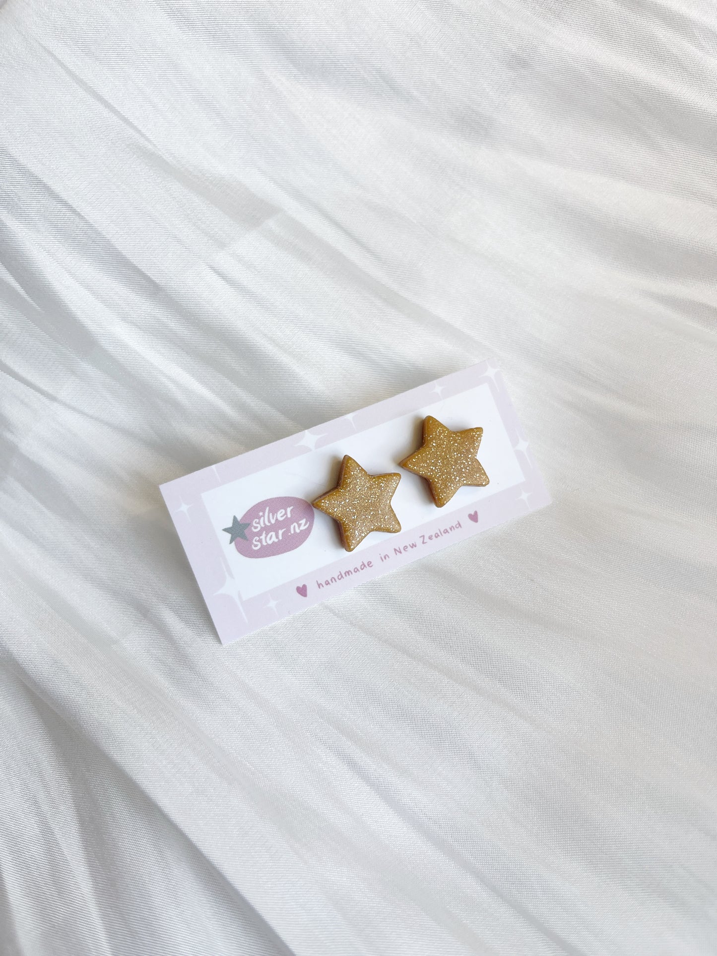 Gold star-shaped earrings from silverstar.nz rest on a light purple card reading handmade in New Zealand, elegantly set against flowing white fabric.