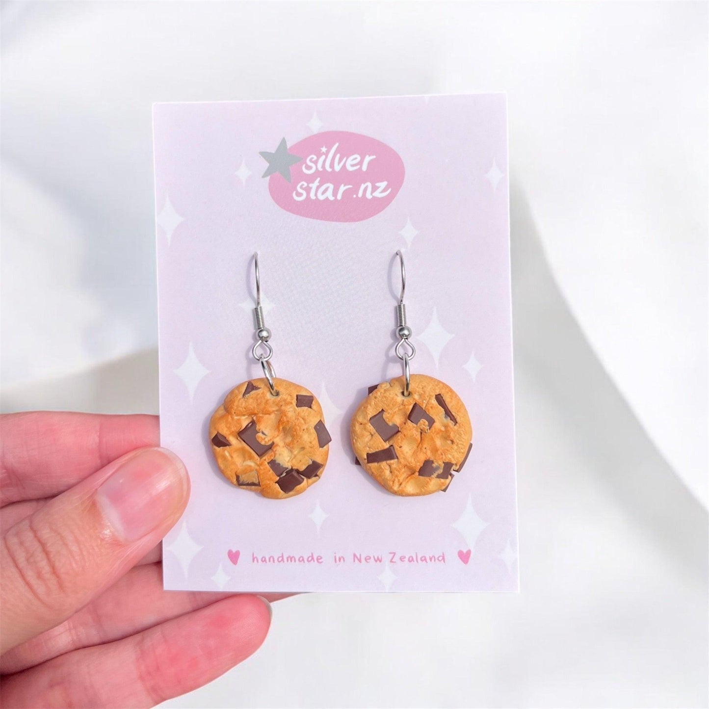 A hand showcases a card featuring the Chocolate Chip Cookie Earrings by silverstar.nz, highlighting their distinctive design as cookie-shaped handmade earrings made from polymer clay. These delightful accessories feature chocolate chip details and proudly declare they are crafted in New Zealand.