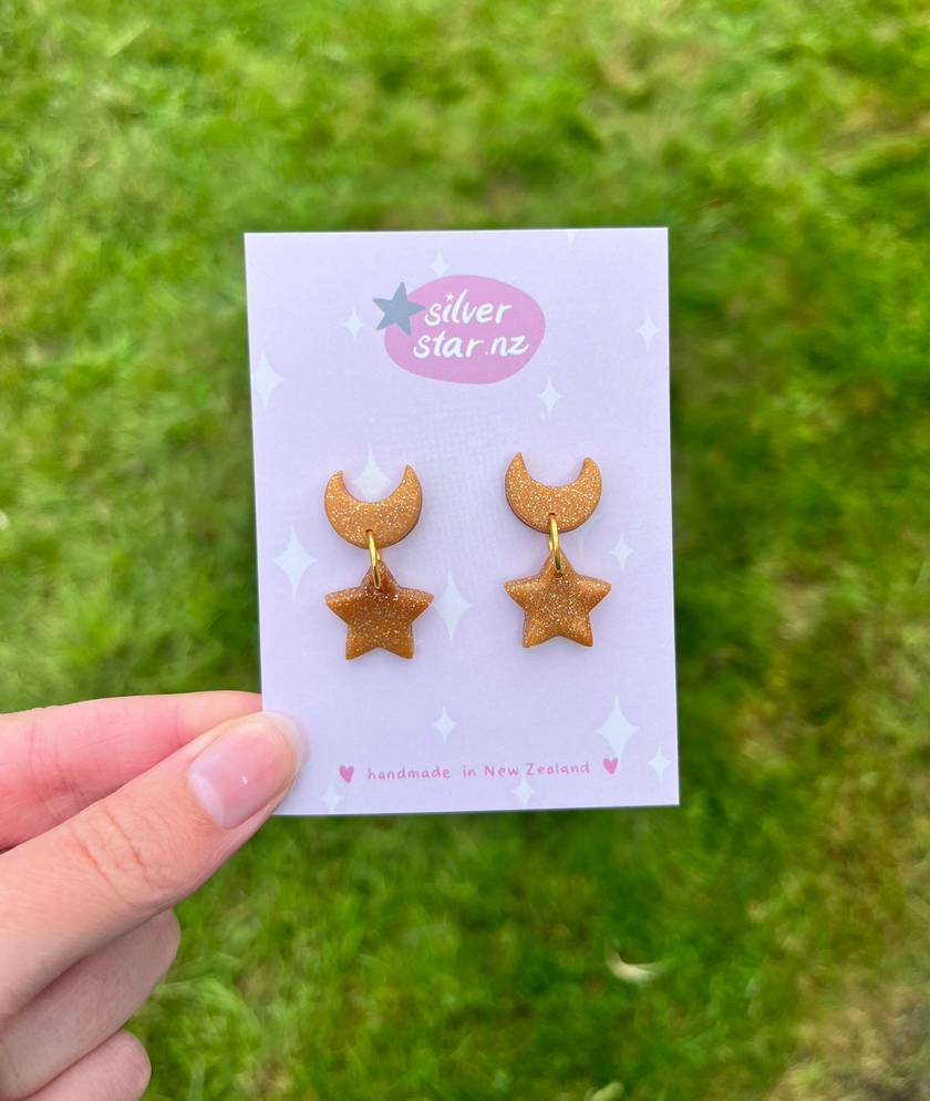 A hand displays a card featuring Star + Moon Polymer Clay Earrings with funky gold glitter designs shaped like crescent moons and stars. The card shows silverstar.nz with stars and diamonds, and handmade in New Zealand. The grassy background enhances these unique earrings beautifully.